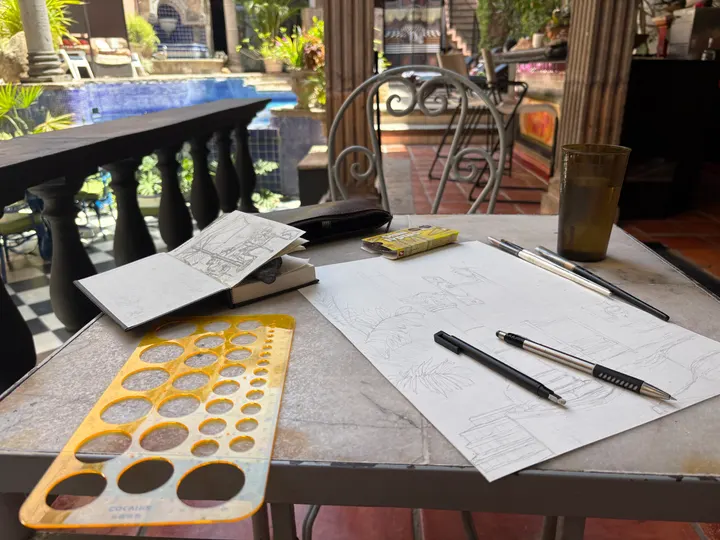 Work in progress: a café table by a pool with a sketchbook, pens, a circle template and a drawing under way