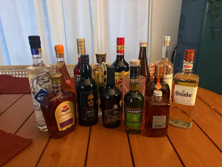 Bottles lined up on a wooden table, among them Seventh Heaven gin, Crown Royal, Campari, Yamazakura whisky, St-Germain, sweet vermouth, Amaro Lucano, Green Chartreuse, Cointreau, Nikka From the Barrel and El Jimador reposado