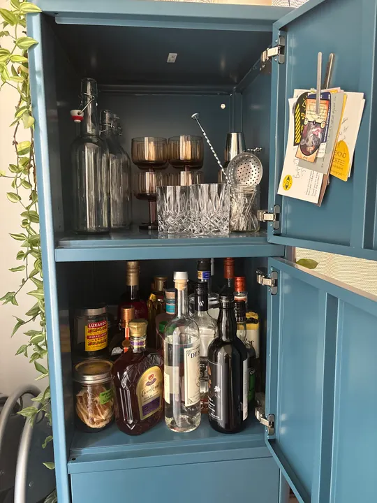 Blue metal bar cabinet with its doors open: amber coupes, cut-glass tumblers, a bar spoon and strainer on the top shelf; bottles, Luxardo cherries and a jar of dried grapefruit below