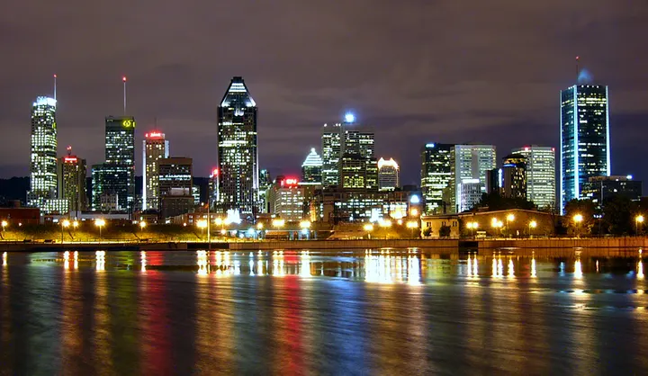 The Montreal skyline at night across the river, its towers lit, the low clouds above glowing a dull purple-orange, coloured reflections on the water.