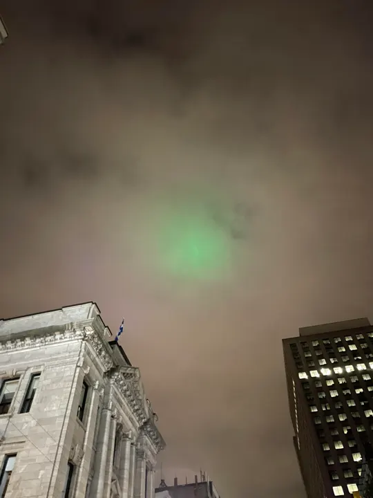 Looking straight up between a stone building flying the Quebec flag and an office tower at night: the low cloud glows orange-brown, with a hazy green spot of light in it.