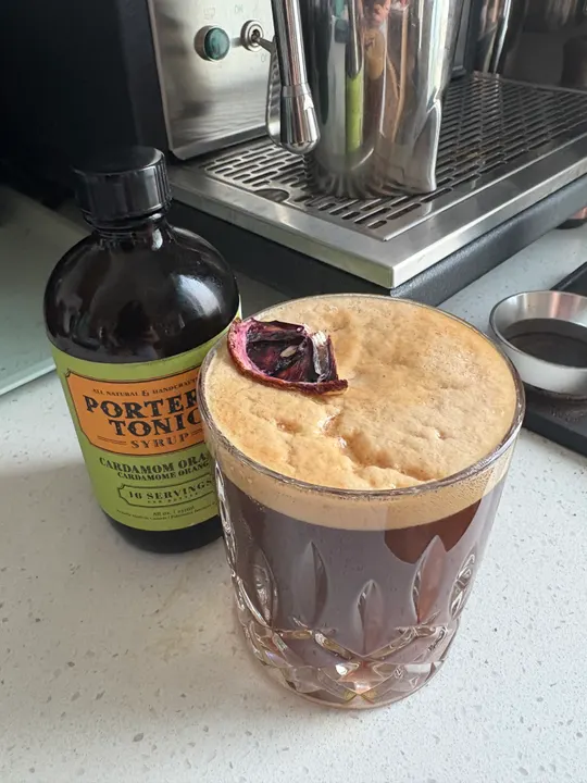 Coffee tonic with a creamy head and a dehydrated blood orange garnish, next to a bottle of Porter’s tonic and an espresso machine
