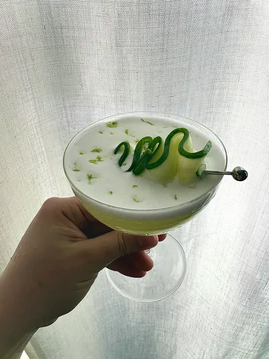 Hand holding a pale green sour in a coupe against a white curtain, a cucumber ribbon on a pick, the foam flecked with lime zest