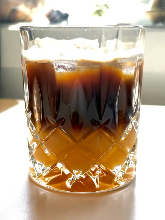 Cut-glass tumbler of orange juice with a dark layer of espresso floating on top, lit by the window