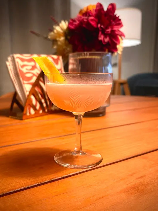 A pale pink sour in an etched coupe on a wooden table, a lemon twist hooked over the rim, with a vase of red and yellow dahlias and a patterned napkin holder behind