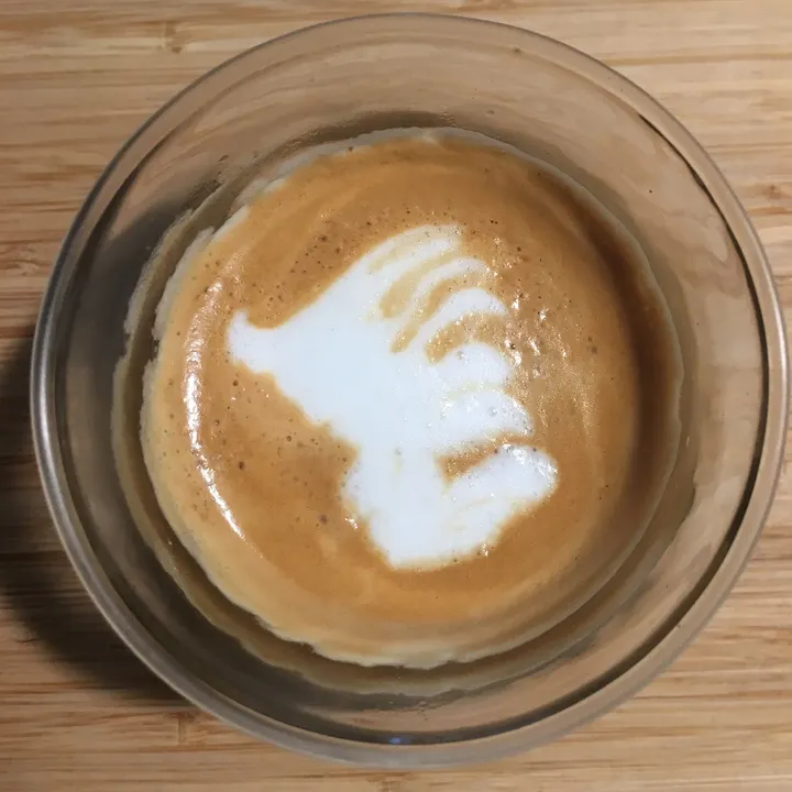 A lopsided white blob on a latte in a glass cup, on a wooden counter