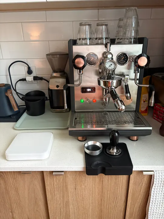 Kitchen counter with a dual-gauge espresso machine, a grinder, a tamper on its mat and a scale