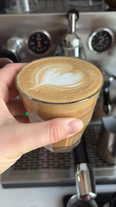 Hand holding a latte in a glass by the machine, a small rosetta on top