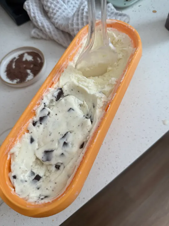 Bay leaf ice cream studded with thin squares of dark chocolate in a long yellow loaf tin, a spoon resting in it